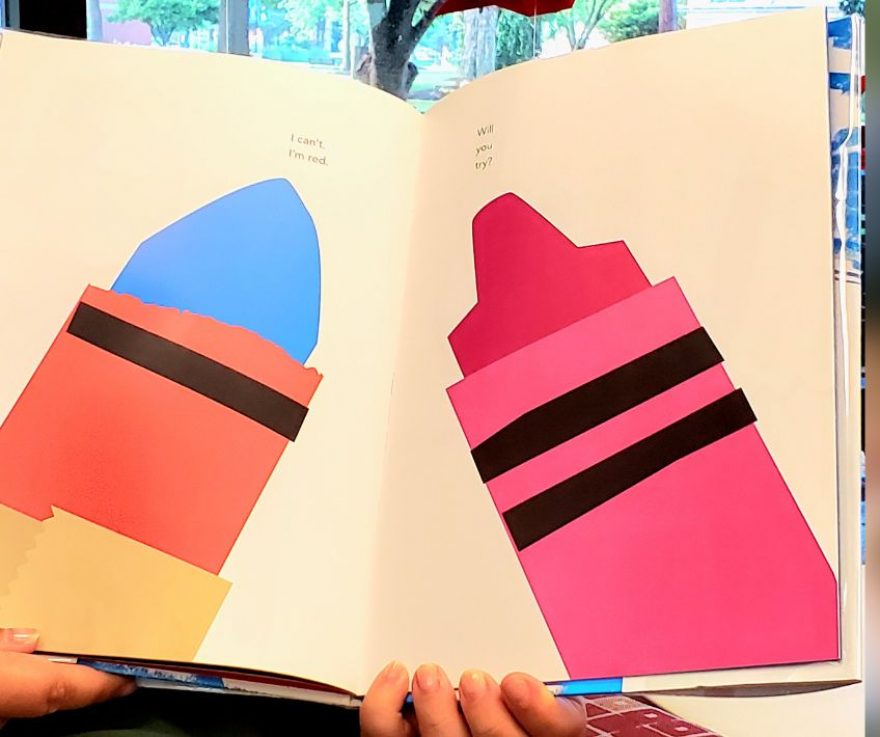 A picture book held open so that two pages are showing. One page has a drawn blue crayon with a red wrapper on it. The other page has a pink crayon on it.