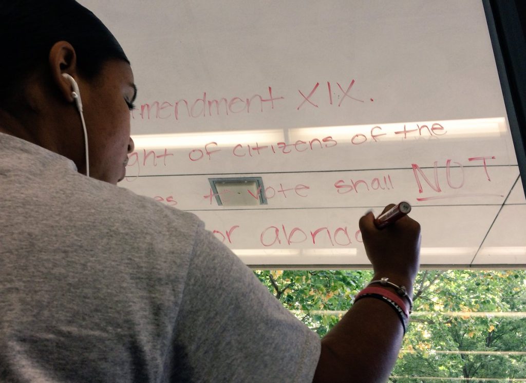 A young person writing out a part of the Constitution on the window. They are wearing a grey shirt and have a white earbud in their ear.