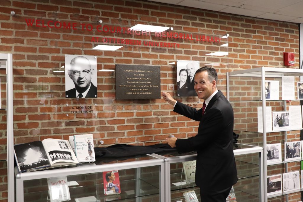 President Comerford standing in front of the new Courtright plaque, a clear wall with photos.