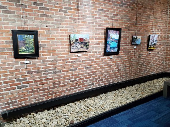 A brick wall with five pieces of art hanging on it. There are stones at the bottom of the wall, then a smaller divider and blue carpet showing.