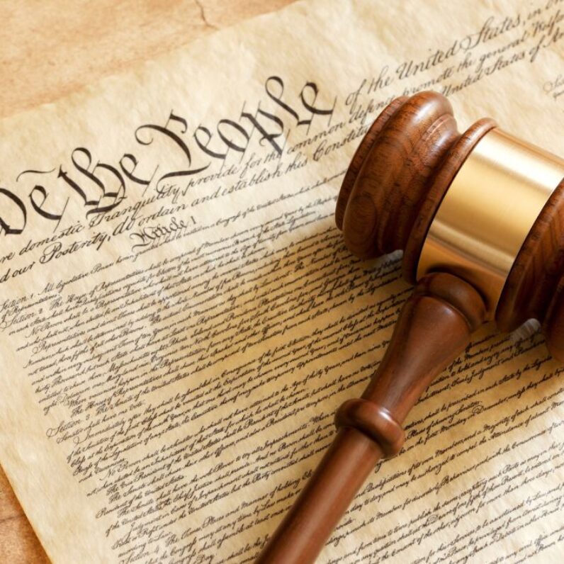 An image of the United States Constitution with a gavel on it. A blurred flag is in the background.