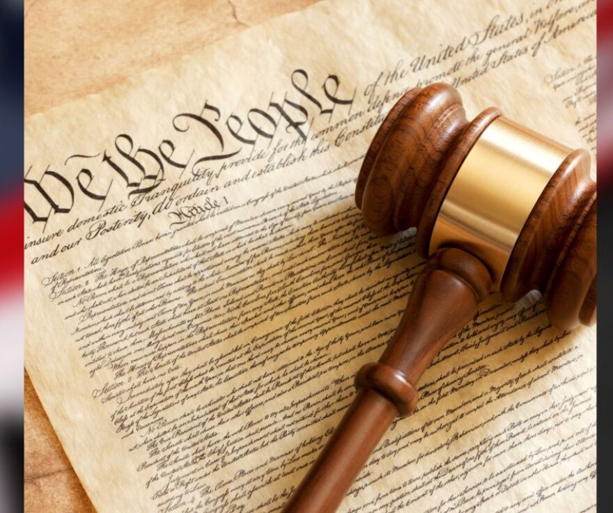 An image of the United States Constitution with a gavel on it. A blurred flag is in the background.