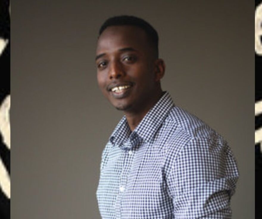An image of Abdi Nor Iftin, a young Black man, smiling at the camera. Behind his photo is a stretched out image of the cover of his book, Call Me American. It is barely visible.