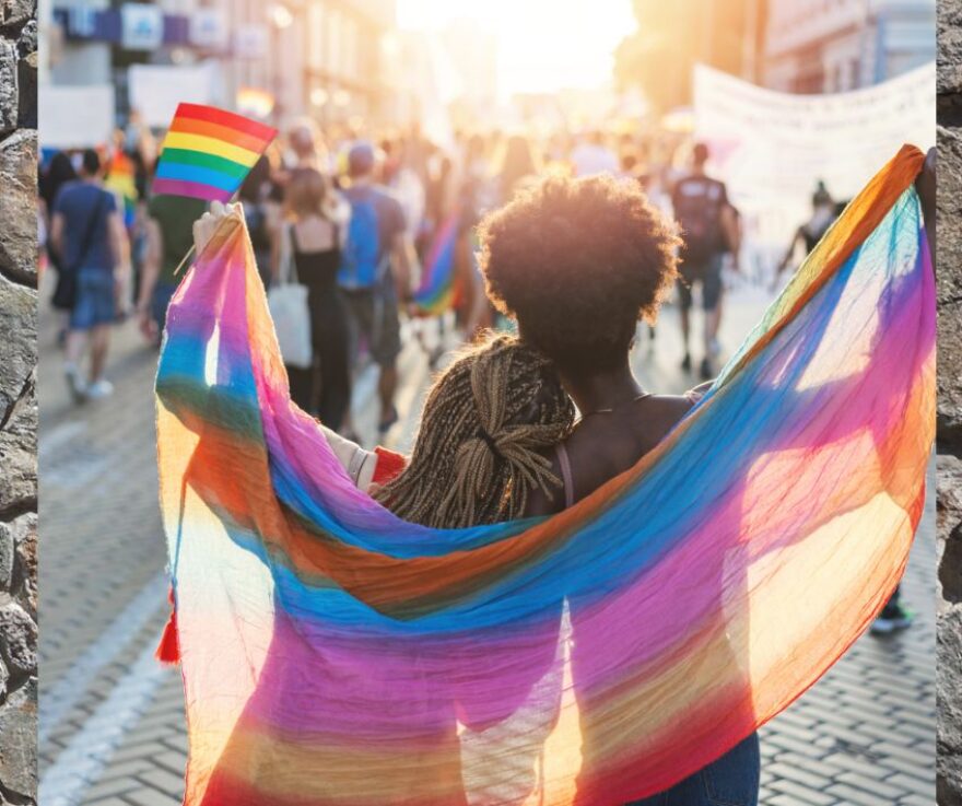 Two people facing away from the camera, holding a queer pride flag over their shoulders.