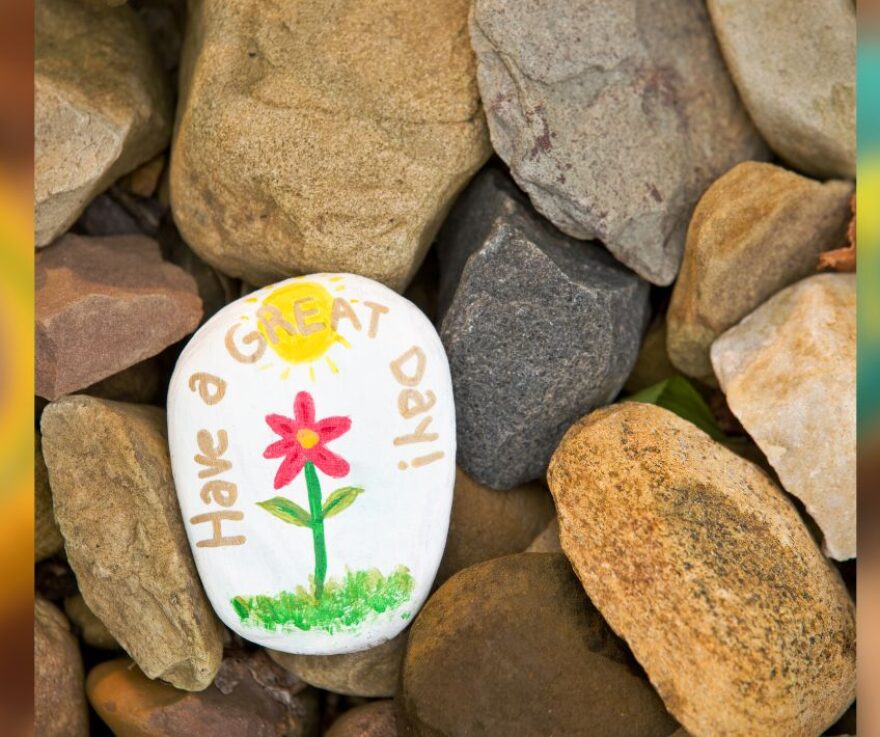 A pile of rocks, with one rock painted in the center. The painted rock has a flower and a sun on it and says Have A Great Day.