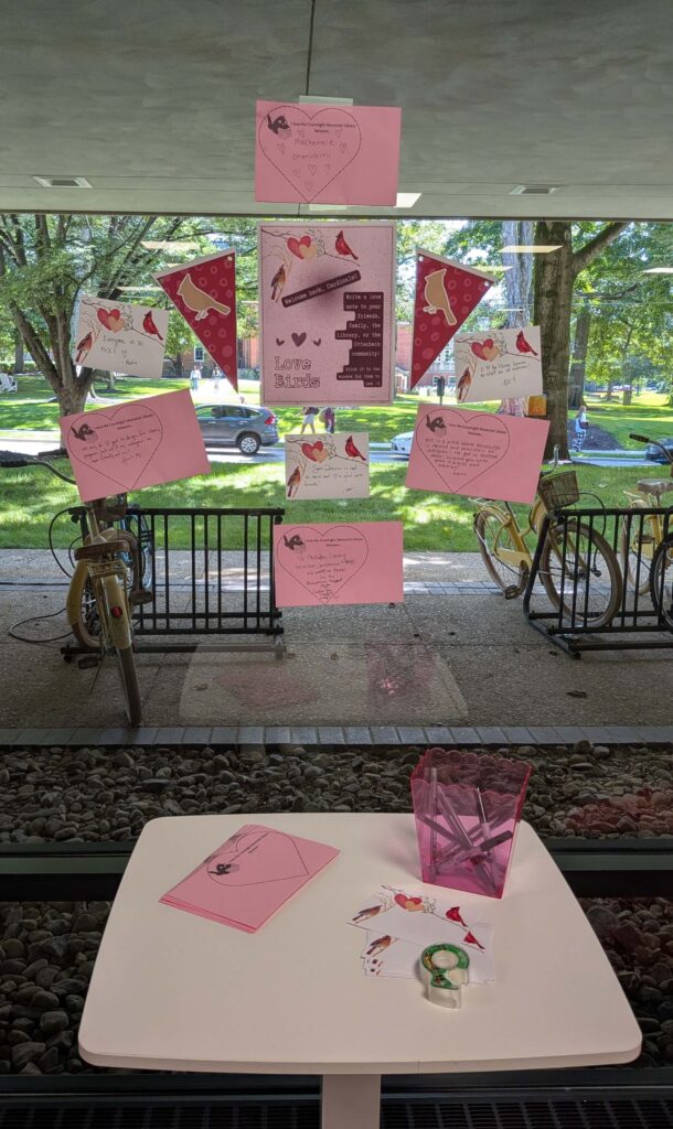 The window wall on the 1st floor of the Courtright Memorial Library, with "Love Bird" activity instructions and some already completed love notes taped to the central window. Image also includes the table sitting in front of the window wall containing love note blanks, tape, and writing utensils.