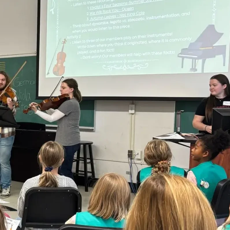 Three students guide a room full of girl scouts through a presentation about music. Two students, a man and a woman playing violas, stand behind a music stand. Two their right, a woman stands behind a computer. The presentation screen behind the presenters reads “How Music is Made.”
