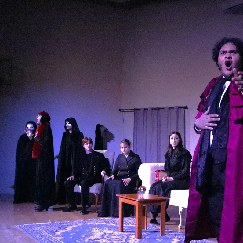 In the foreground a Black man in a red trench coat and 19th century suit belts on a darkened stage, one hand rested on his stomach and the other stretched out behind him. Behind him, three white people, one man and two women, sit on antique furniture with worried expressions. In the background, a woman in a black fur coat with a match red fur hat and scarf sings as she is encircled by three figures in white masks and black, hooded cloaks.